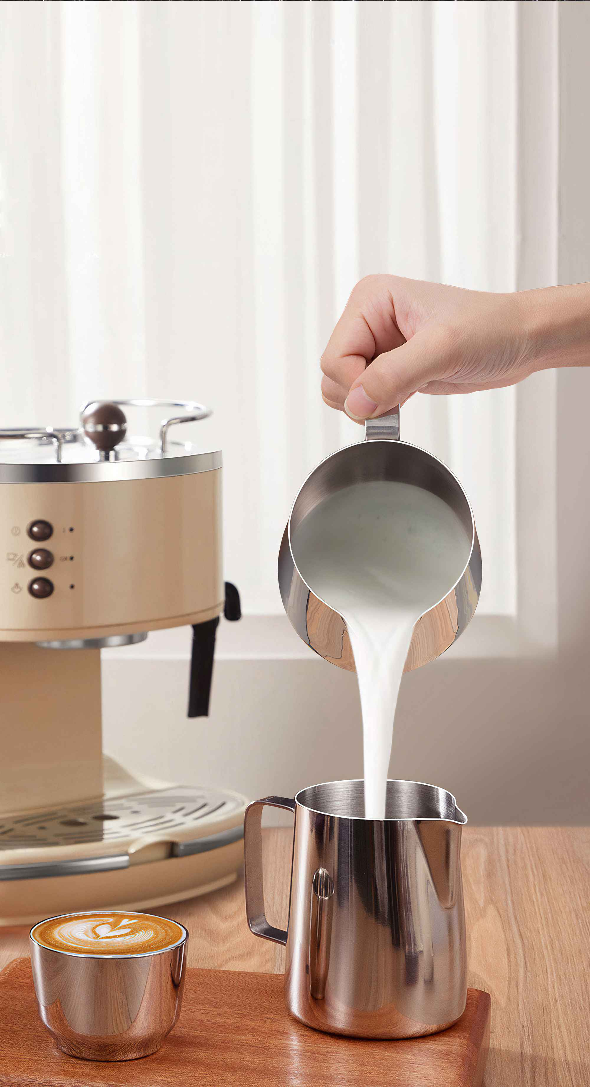 Eine Hand gießt Milch aus einem Krug in einen anderen. Ein Latte steht in der Nähe. Eine Kaffeemaschine ist dahinter.