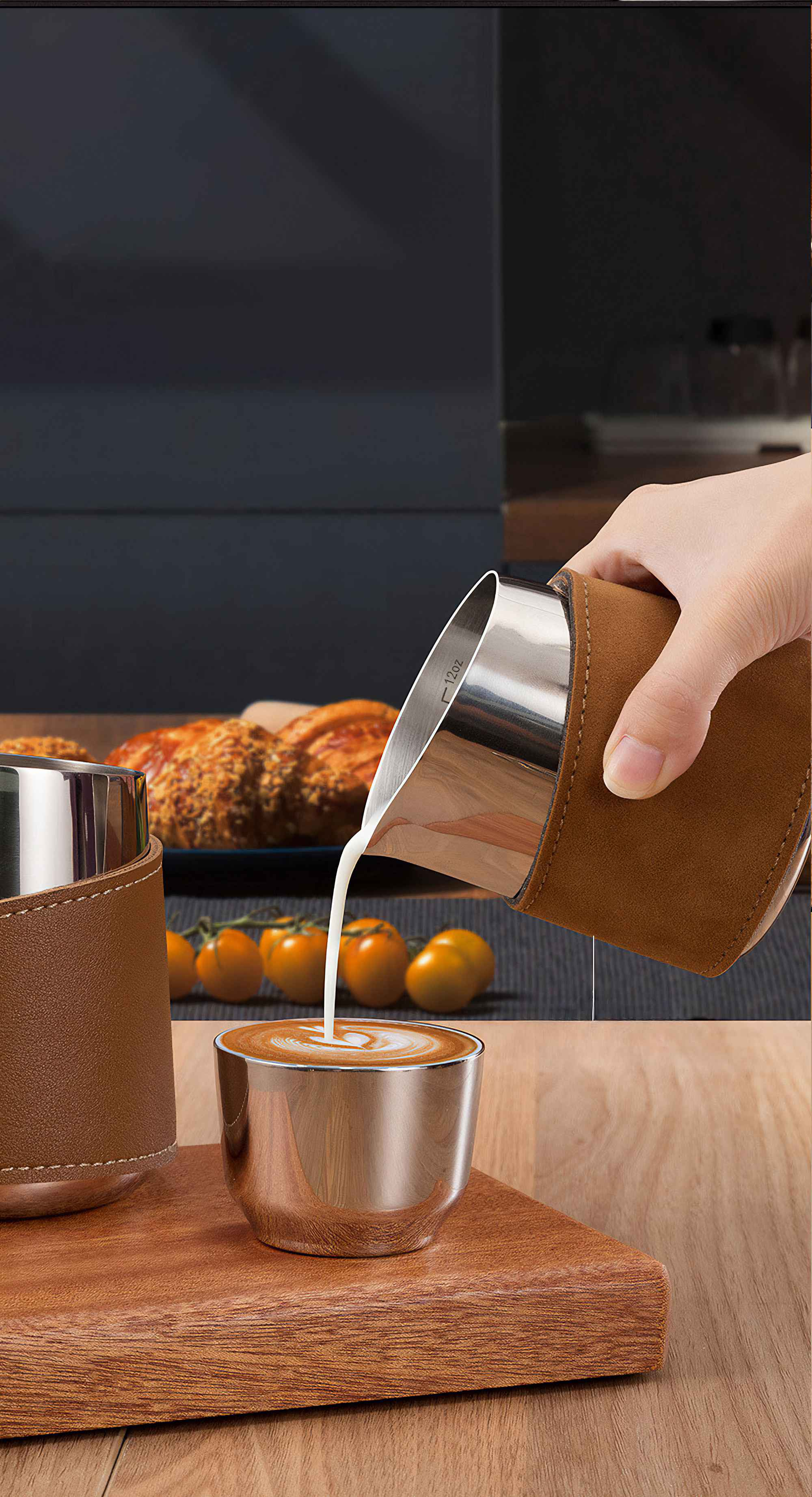Eine Hand gießt Milch in eine Kaffeetasse. Tasse und Milchkanne sind aus Edelstahl mit braunem Leder. Tomaten und Brot sind im Hintergrund.