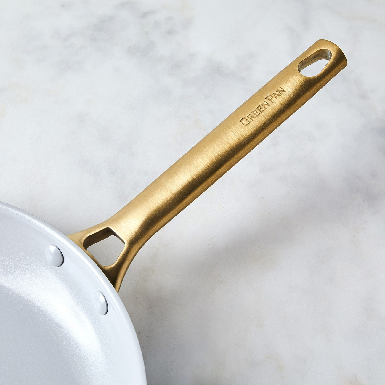 Nahaufnahme eines goldfarbenen Bratpfannengriffs mit der Gravur "GREENPAN". Die Pfanne hat einen weißen Innenraum und liegt auf einer Marmoroberfläche.