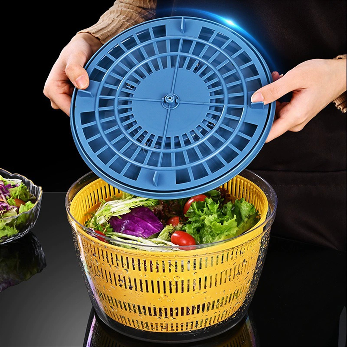 Hände halten einen blauen Salatschleuderdeckel über einem gelben Korb mit Salat und Tomaten. Eine Schüssel Salat steht daneben.