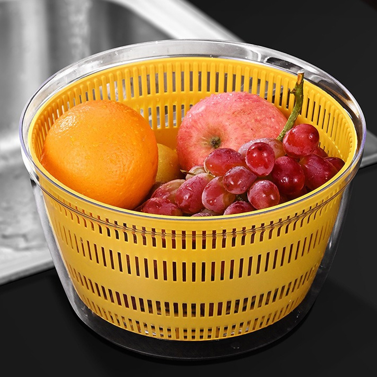 Ein gelber Fruchtspinner enthält eine Orange, einen roten Apfel, Trauben und eine Zitrone. Der Spinner steht in einer klaren Schüssel auf schwarzem Grund.