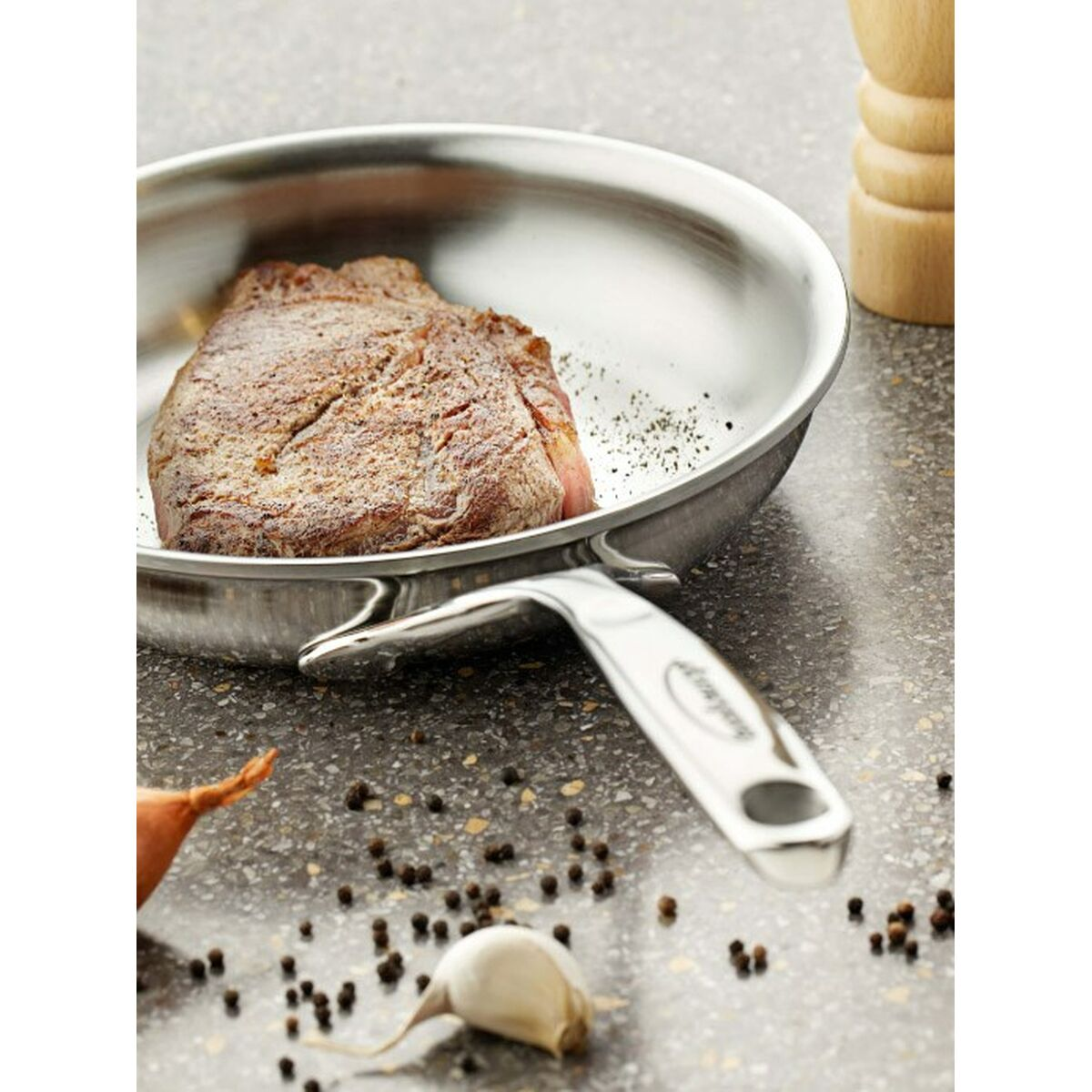 Un steak assaisonné dans une poêle en acier inoxydable. Des grains de poivre, de l'ail et de l'oignon dispersés sur un comptoir moucheté. Un moulin à poivre en bois à proximité.