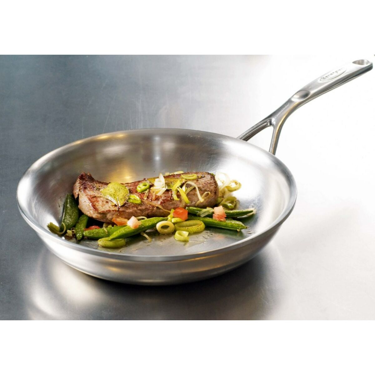Une poêle en acier inoxydable avec un steak cuit et des légumes. Le steak est assaisonné et garni. La poêle est sur une surface métallique.