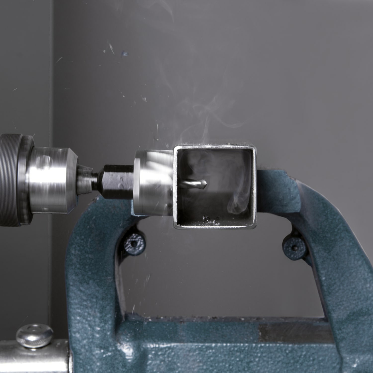 Metalowa rura jest zaciśnięta. Wiertło tworzy otwór. Dym unosi się. Szare tło. Niebieski zacisk trzyma rurę.