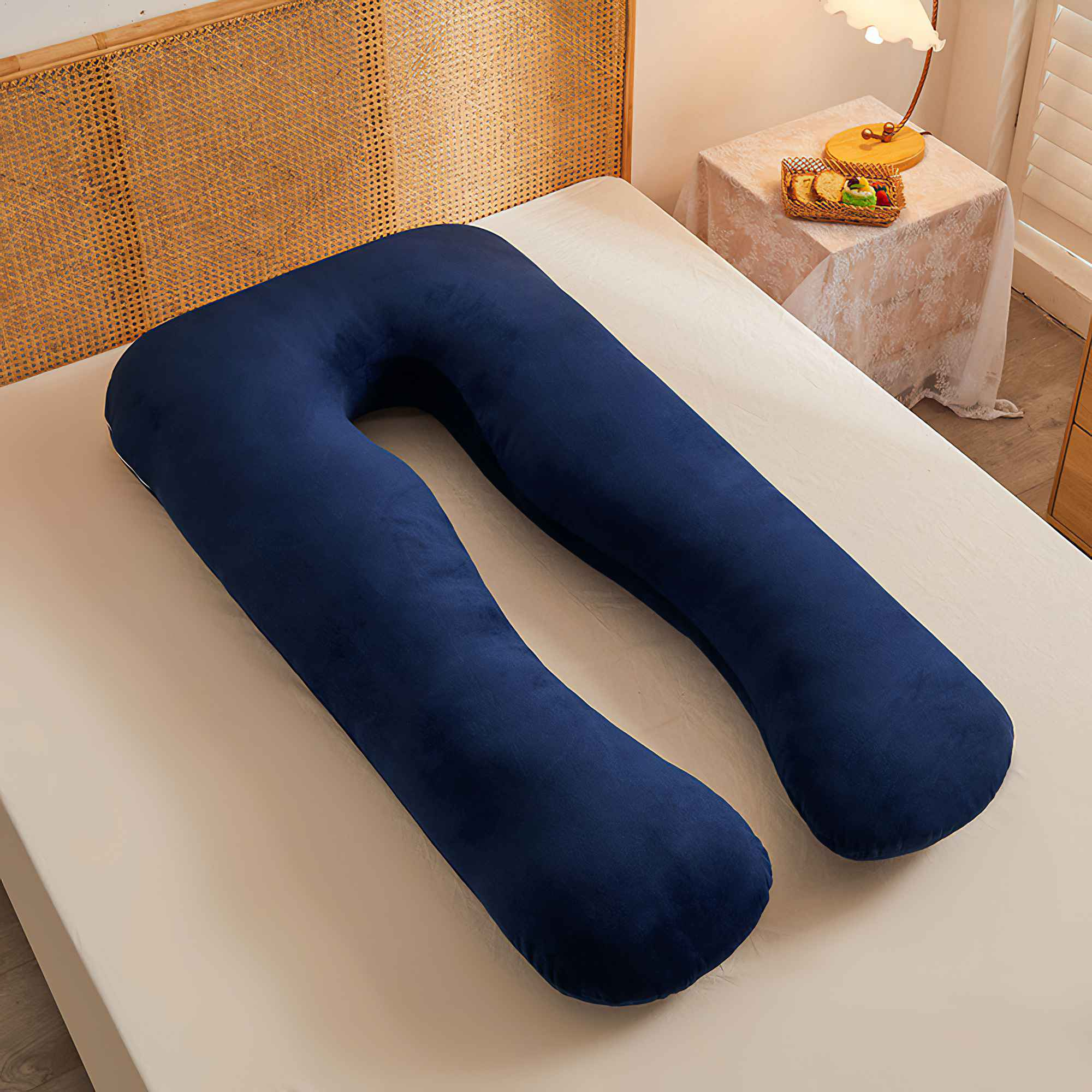 Ein marineblaues U-förmiges Schwangerschaftskissen liegt auf einem Bett. Daneben steht ein Beistelltisch mit Lampe und einem Korb mit Essen.