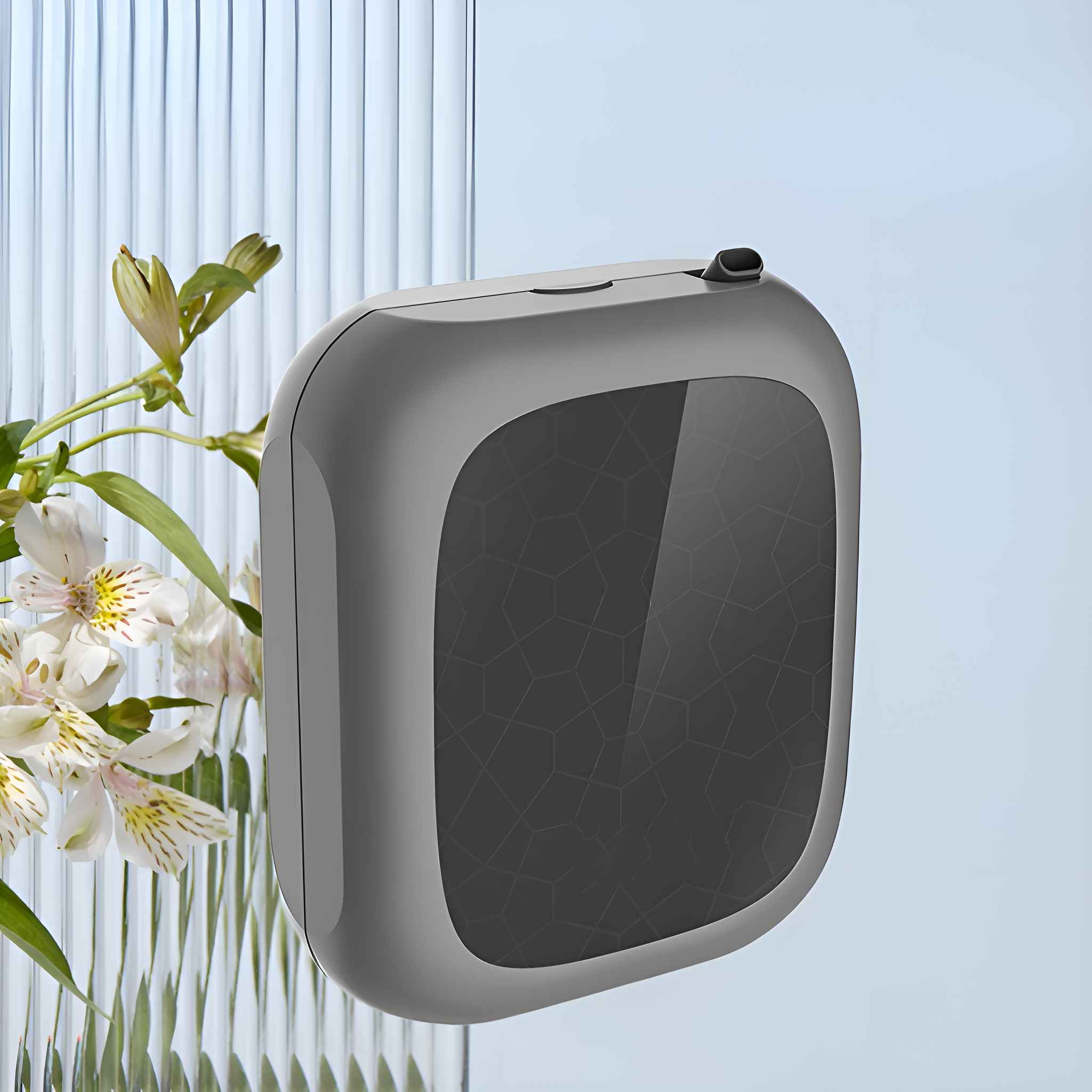 Ein graues, quadratisches Gerät mit gemusterter Vorderseite, weiße Blumen in einer Vase und ein hellblauer Hintergrund.