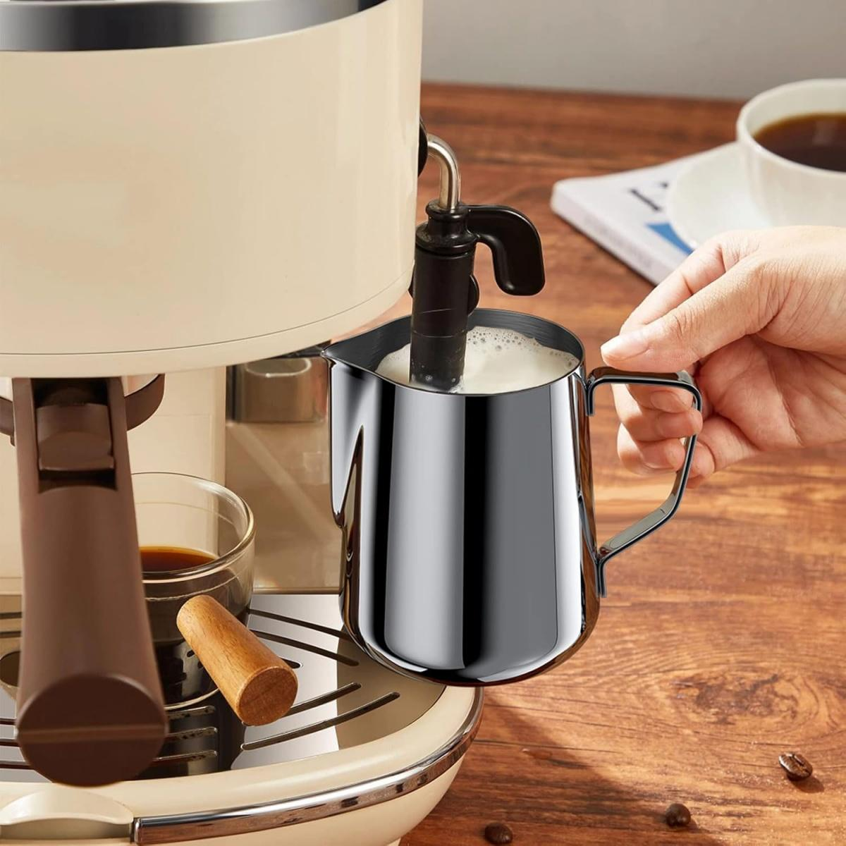 Eine Hand hält einen Stahlkrug mit geschäumter Milch in der Nähe einer Espressomaschine. Kaffeetasse und Holzoberfläche sind sichtbar.