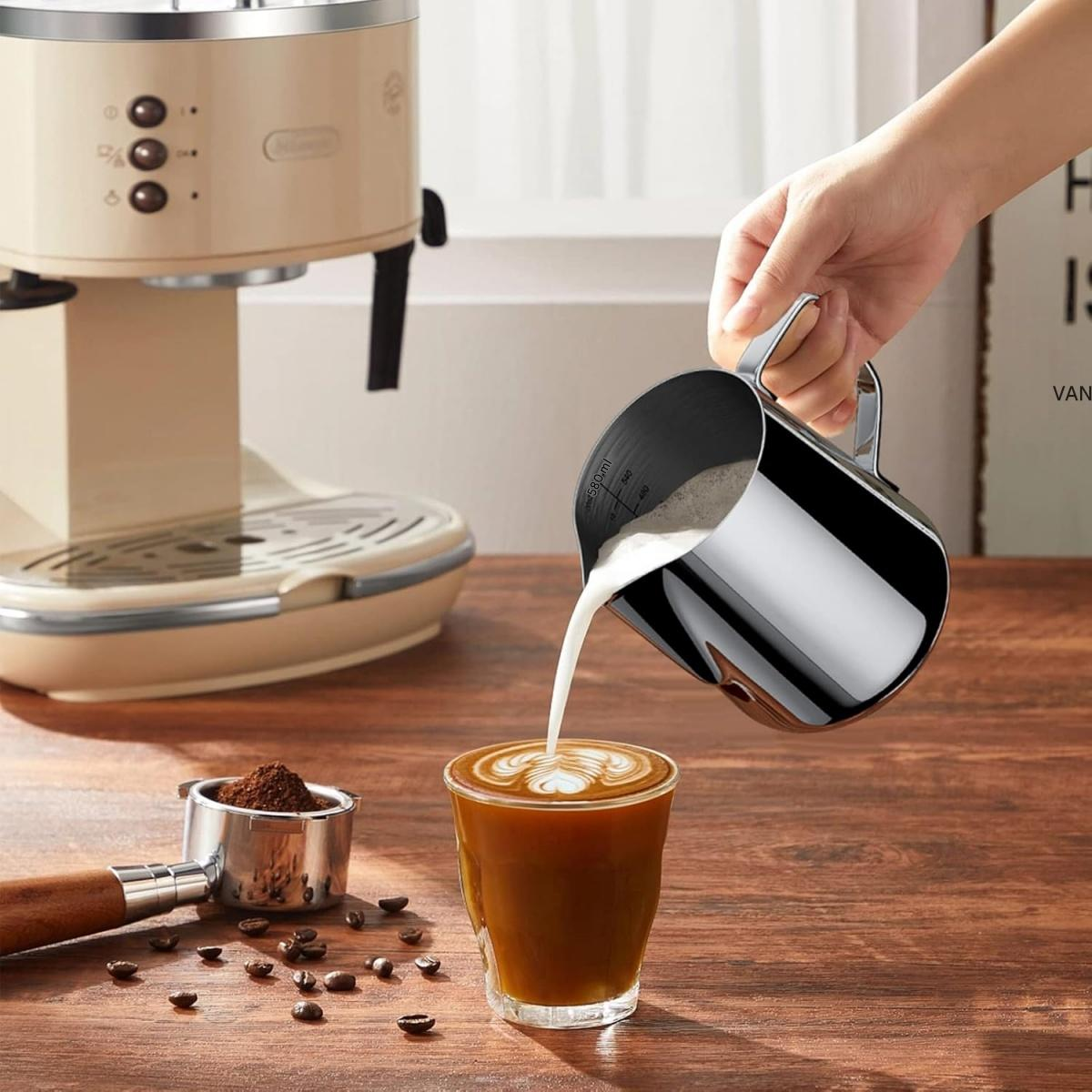 Eine Hand gießt Milch in eine Kaffeetasse und kreiert Latte Art. Kaffeemaschine, Siebträger und Bohnen sind ebenfalls zu sehen.