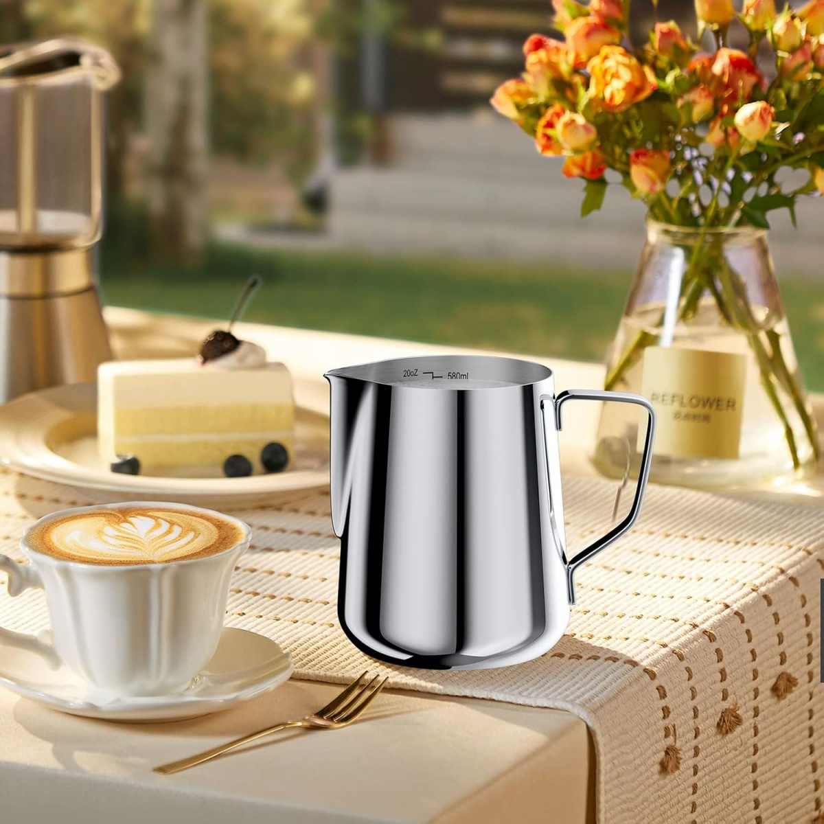 Ein Tischarrangement umfasst eine Kaffeetasse, Kuchen und einen Edelstahlkrug. Im Hintergrund steht eine Vase mit Blumen.