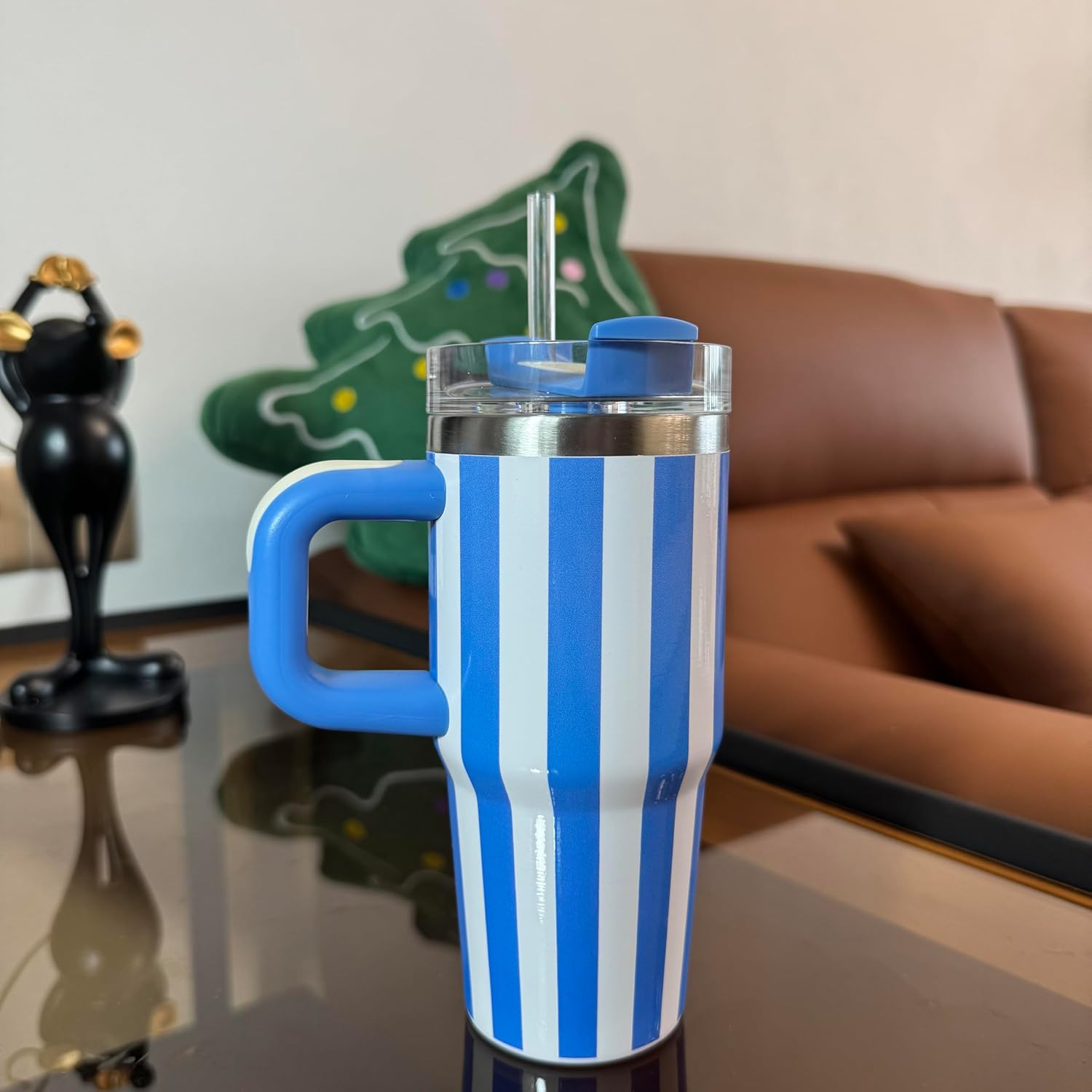 Ein blau-weiß gestreifter Becher mit blauem Griff und Deckel auf einer reflektierenden Oberfläche. Im Hintergrund ein grünes, baumförmiges Kissen.