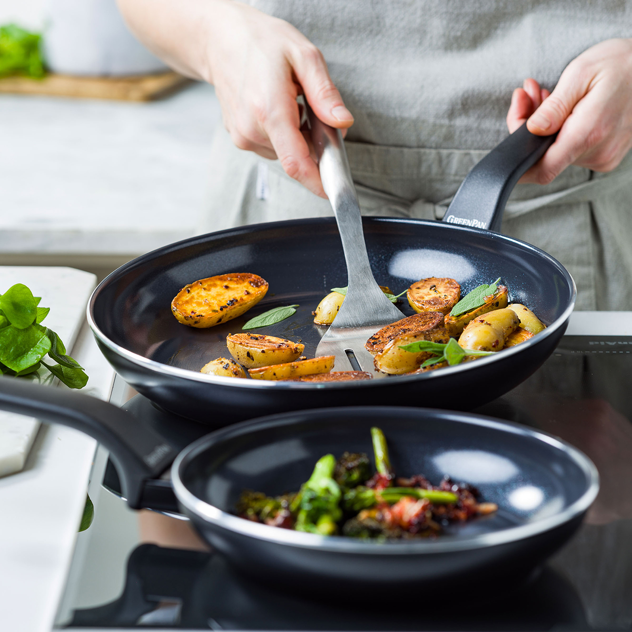 Eine Person kocht Essen in zwei Pfannen. Eine Pfanne hat Kartoffeln, die andere Gemüse. Die Person benutzt einen Spatel.
