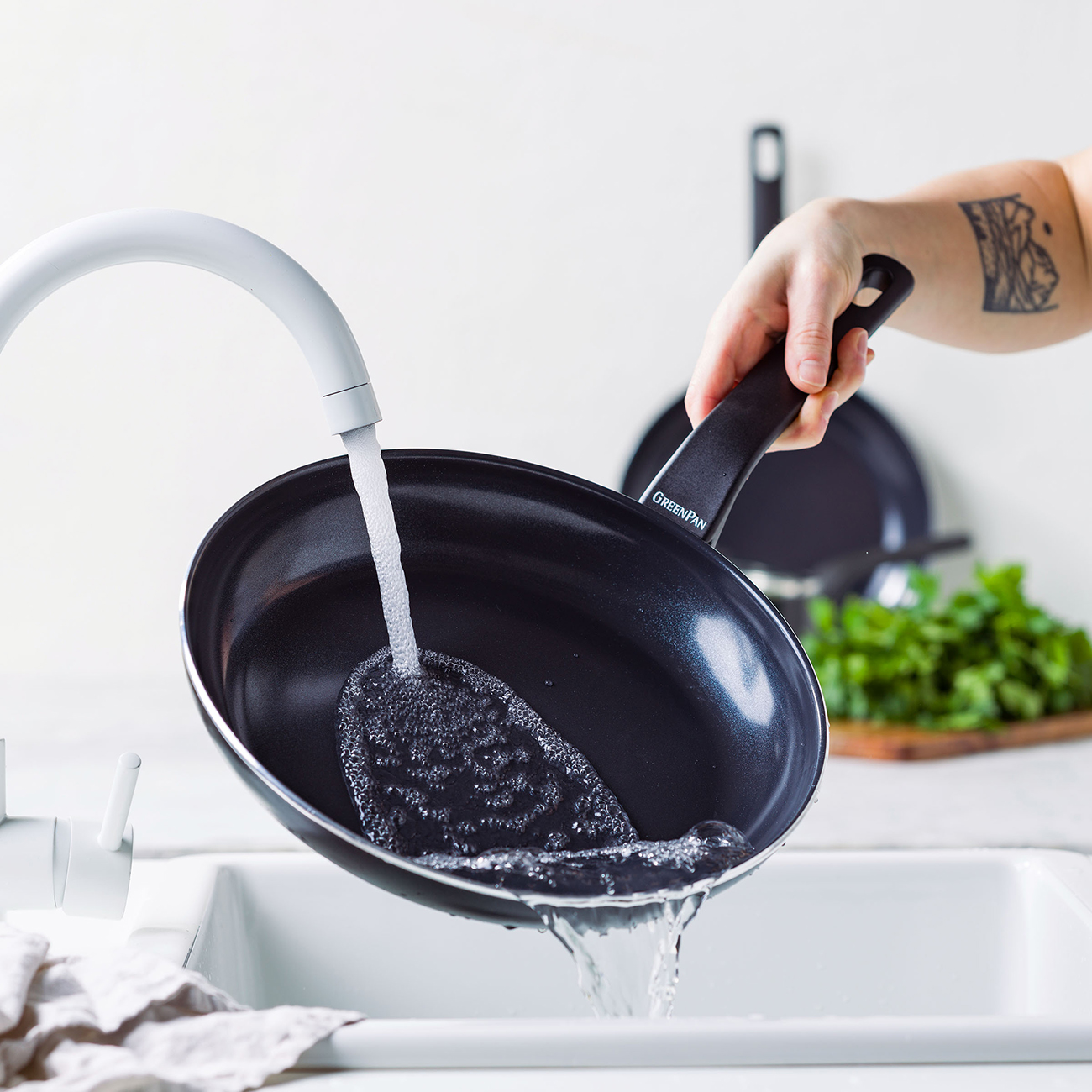 Eine Person wäscht eine schwarze Bratpfanne unter einem weißen Wasserhahn. Wasser gießt in die Pfanne. Andere Pfannen und Kräuter sind im Hintergrund.