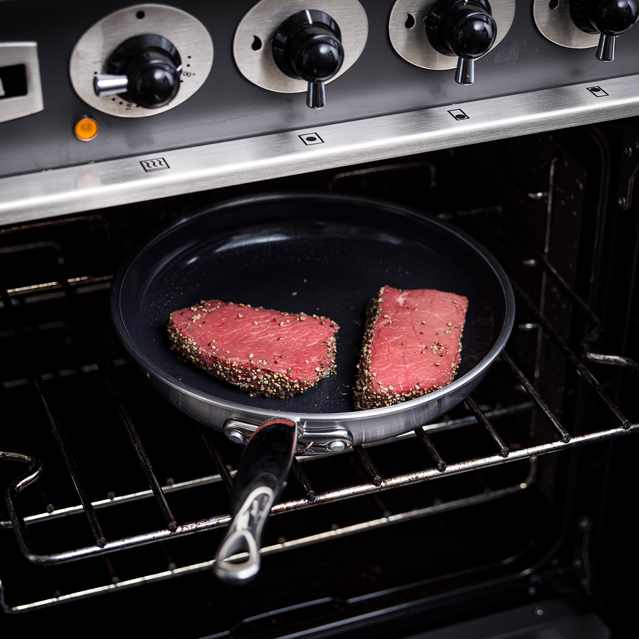 Zwei gewürzte Steaks in einer Pfanne im Ofen. Ofenbedienelemente sind sichtbar. Die Steaks sind rosa und gepfeffert.