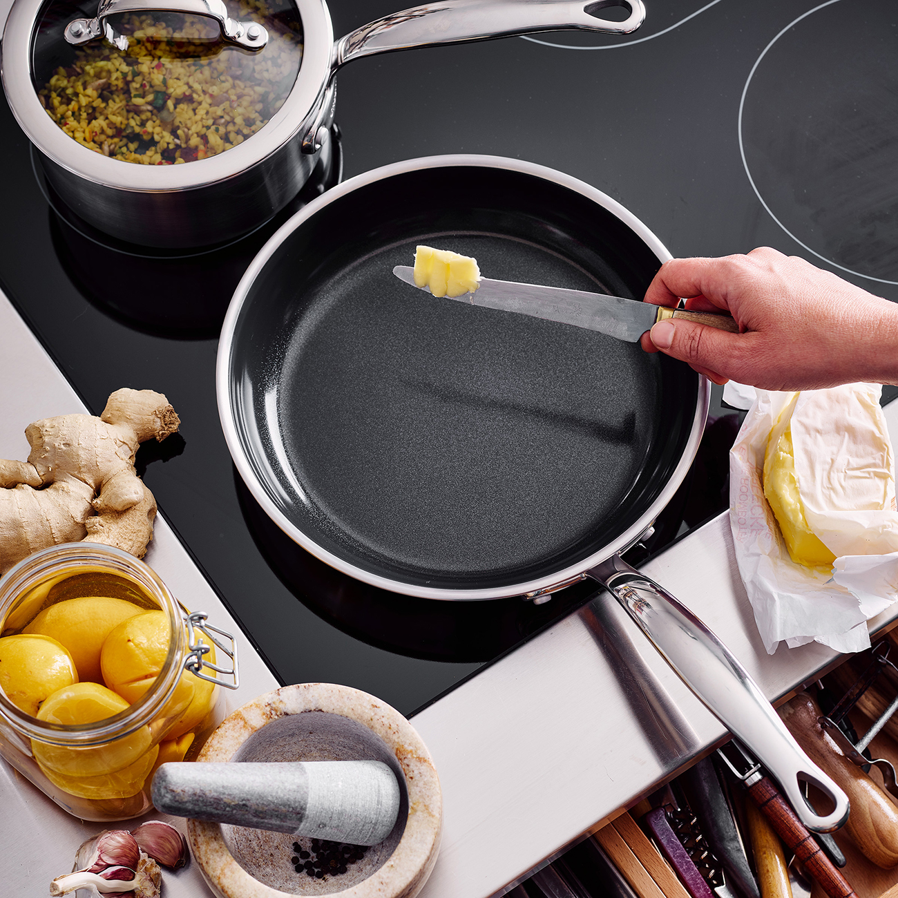 Eine Hand hält ein Messer mit Butter über einer Pfanne. Weitere Gegenstände sind ein Topf mit Essen, Zitronen, Knoblauch, Ingwer und ein Mörser.