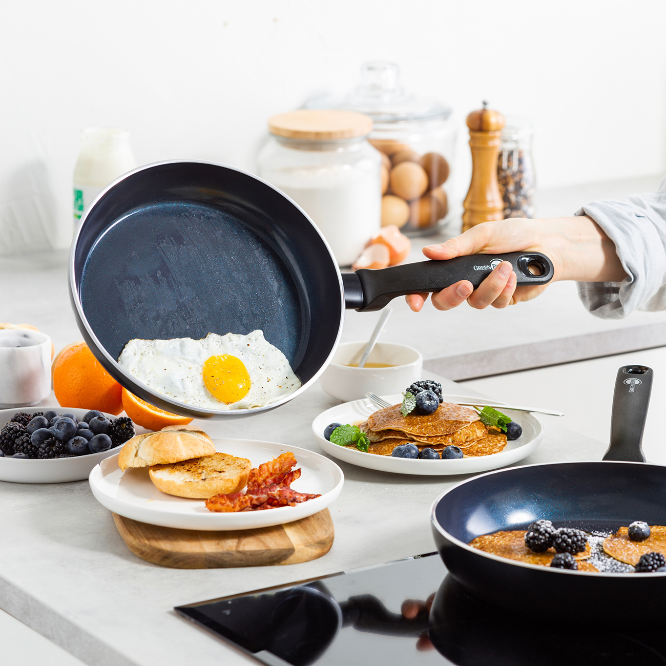 Eine Person hält eine Pfanne mit einem Spiegelei. Auf anderen Tellern liegen Pfannkuchen, Speck und Beeren. Küchenumgebung mit Essen und Zutaten.