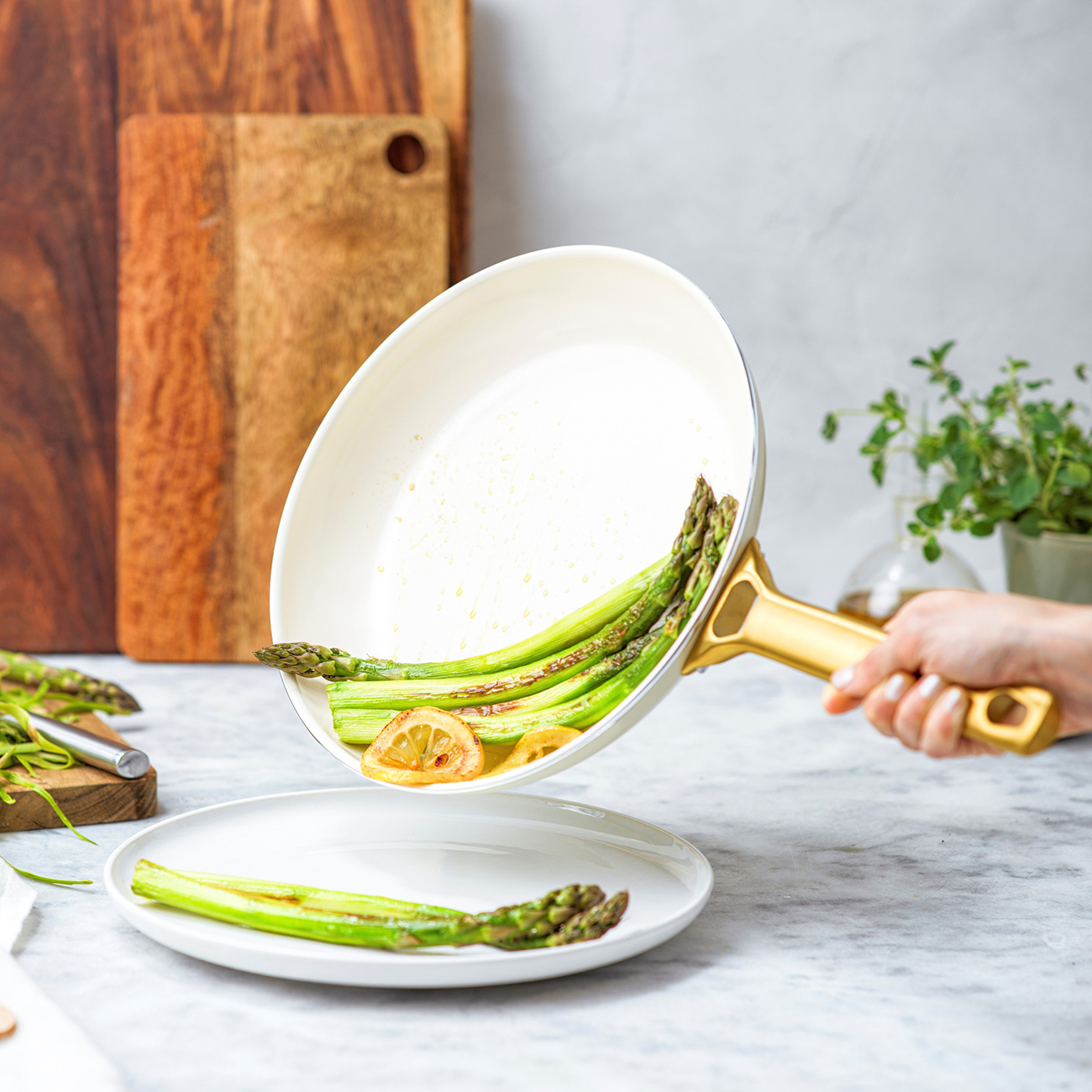 Eine Hand hält eine Pfanne und gießt gekochten Spargel und Zitronenscheiben auf einen weißen Teller. Holzschneidebretter im Hintergrund.