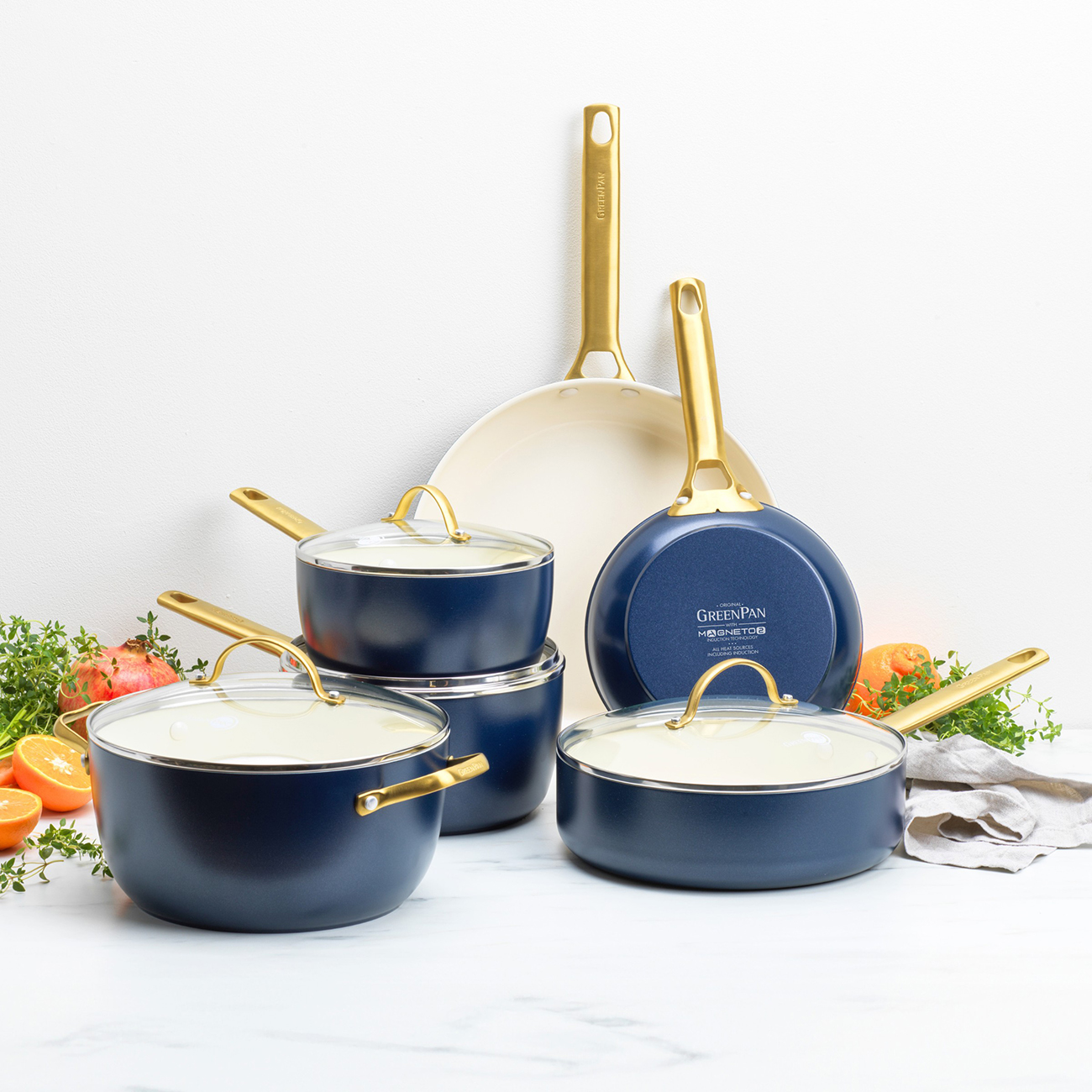 Ein Set aus marineblauem und goldenem Kochgeschirr, einschließlich Töpfen und Pfannen, auf einer weißen Oberfläche. Obst und Kräuter sind um das Kochgeschirr herum platziert.