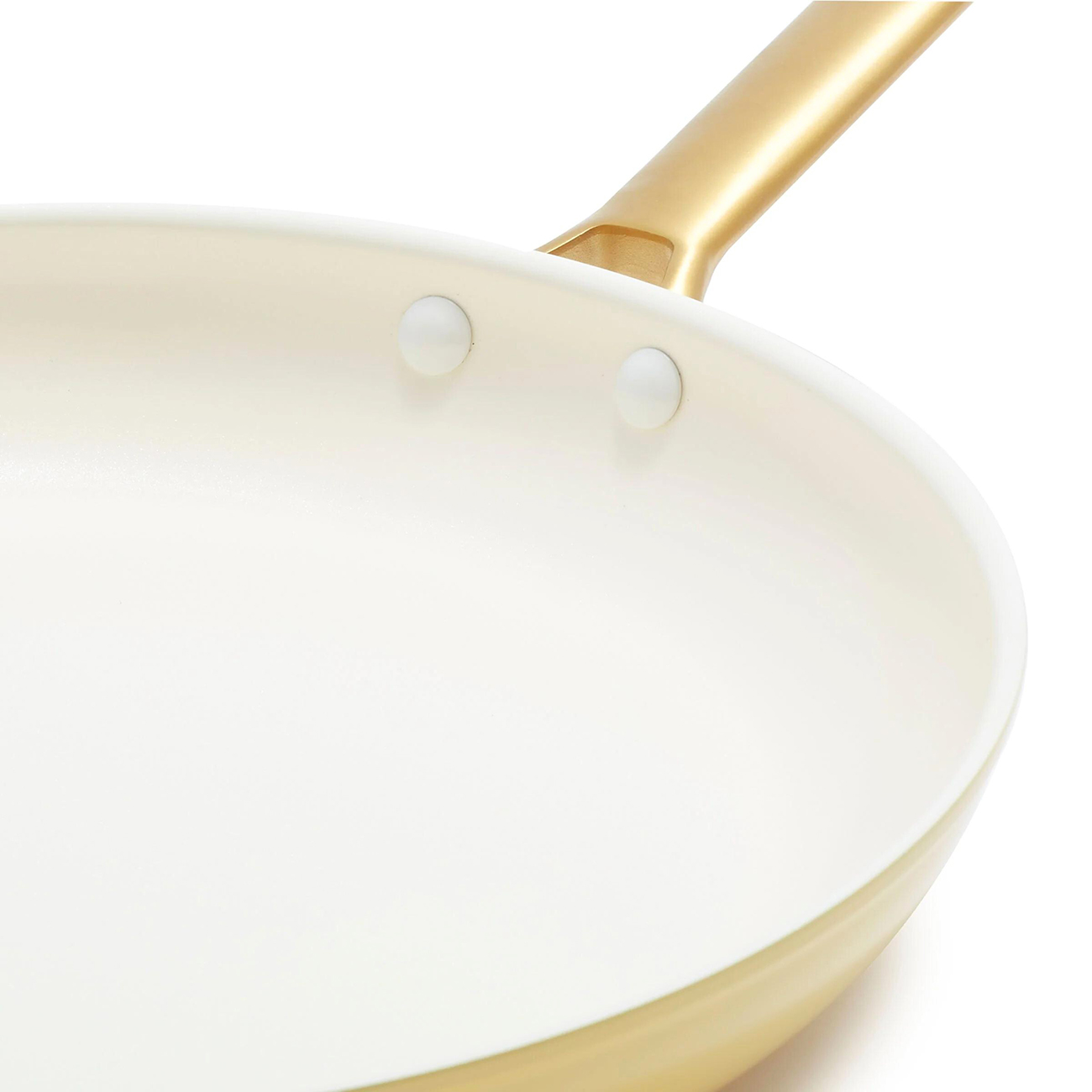 Nahaufnahme einer cremefarbenen Bratpfanne mit goldenem Griff. Zwei weiße Nieten sind sichtbar. Der Hintergrund ist weiß.