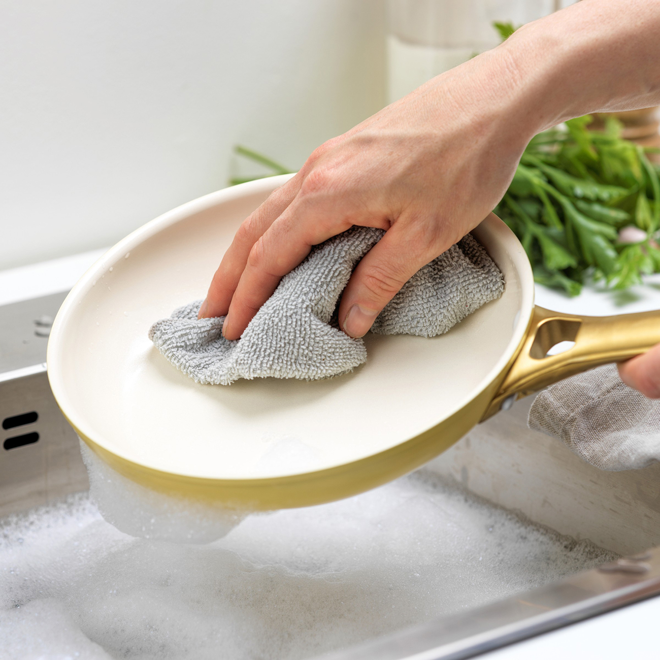 Eine Person wäscht eine Pfanne in einem Waschbecken mit einem grauen Tuch. Die Pfanne ist cremefarben mit einem goldenen Griff. Seifenschaum umgibt die Pfanne.