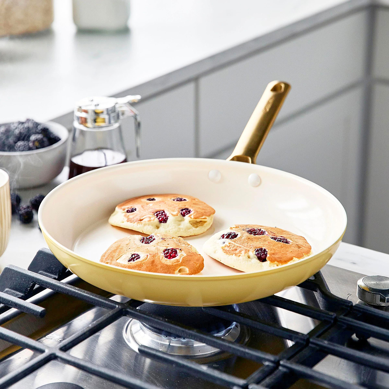 Drei Pfannkuchen mit Beeren in einer gelben Pfanne auf einem Herd. Eine Schüssel mit Brombeeren und Sirup ist im Hintergrund.