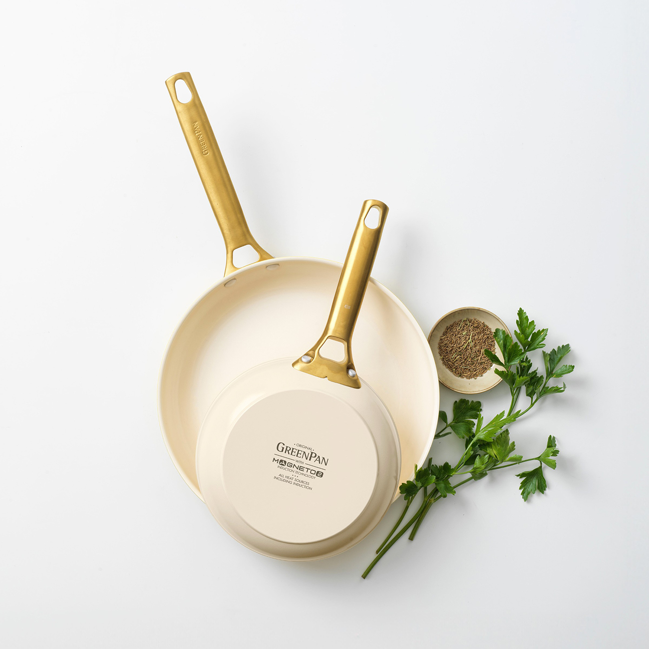 Zwei cremefarbene Pfannen mit goldenen Griffen, eine davon umgedreht, neben einer kleinen Schüssel mit Samen und Petersilie auf weißem Untergrund.