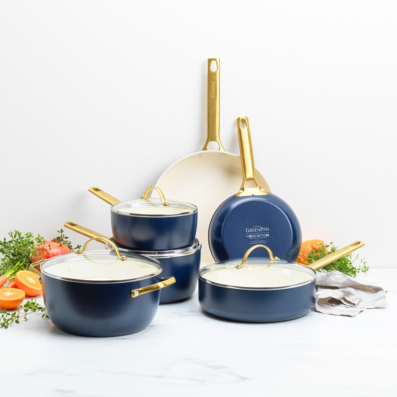 Satz blaue Kochgeschirre mit goldenen Griffen. Enthält Töpfe, Pfannen und Deckel. Karotten, Orangen und Kräuter sind um das Kochgeschirr angeordnet.