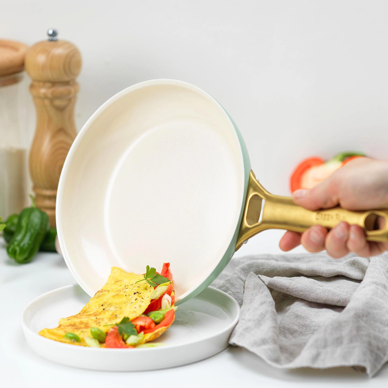 Eine Hand hält eine Pfanne und gießt ein Omelett mit Tomaten und Kräutern auf einen Teller. Die Pfanne ist grün und goldfarben. Weitere Zutaten sind sichtbar.