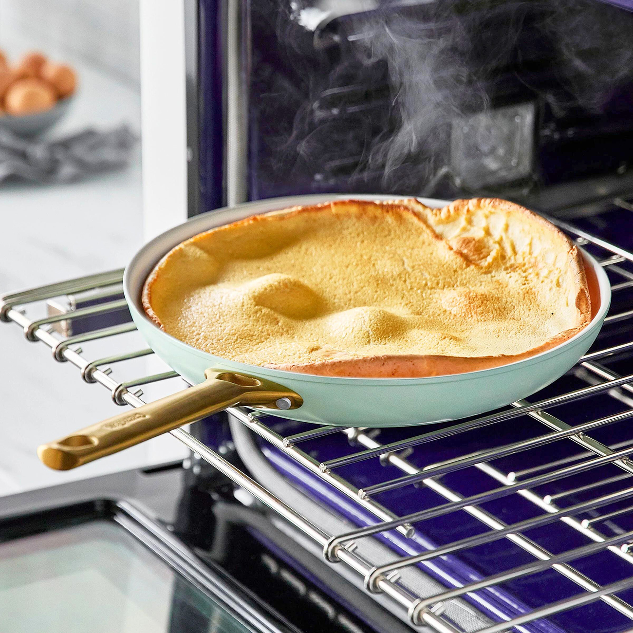 Ein Dutch-Baby-Pfannkuchen in einer Pfanne im Ofen. Dampf steigt aus dem Pfannkuchen auf. Die Pfanne steht auf einem Rost, im Hintergrund Eier.