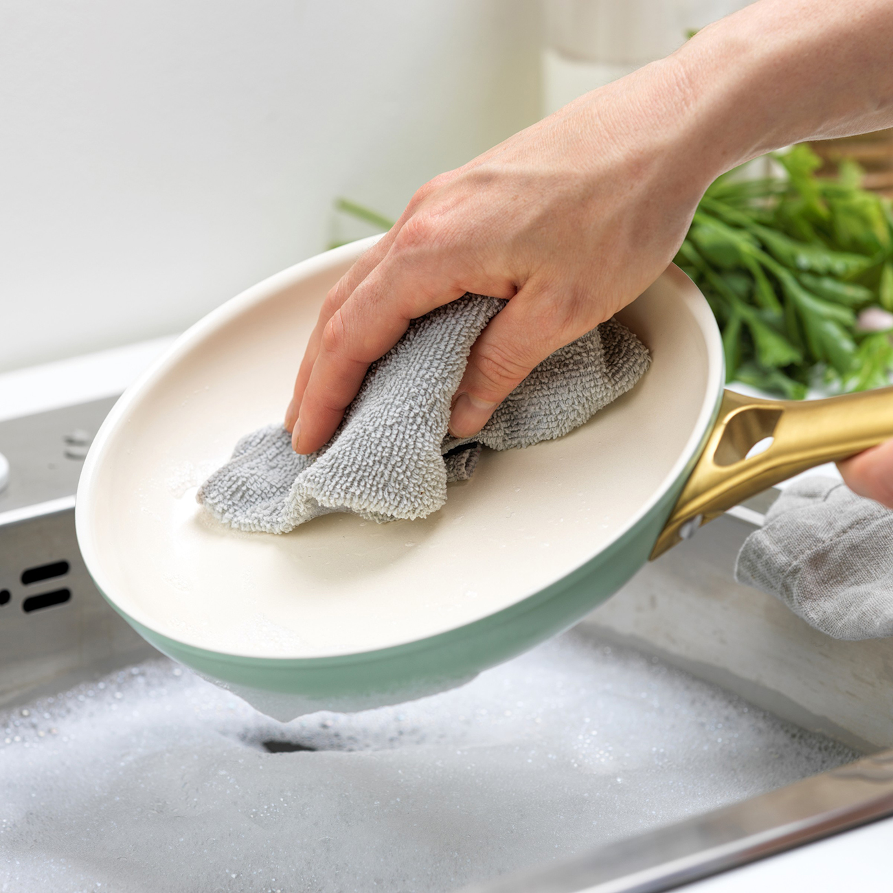 Eine Person wäscht eine Pfanne in einem mit Seifenwasser gefüllten Waschbecken. Die Pfanne ist grün und beige. Eine Hand hält ein graues Tuch, um die Pfanne abzuwischen.