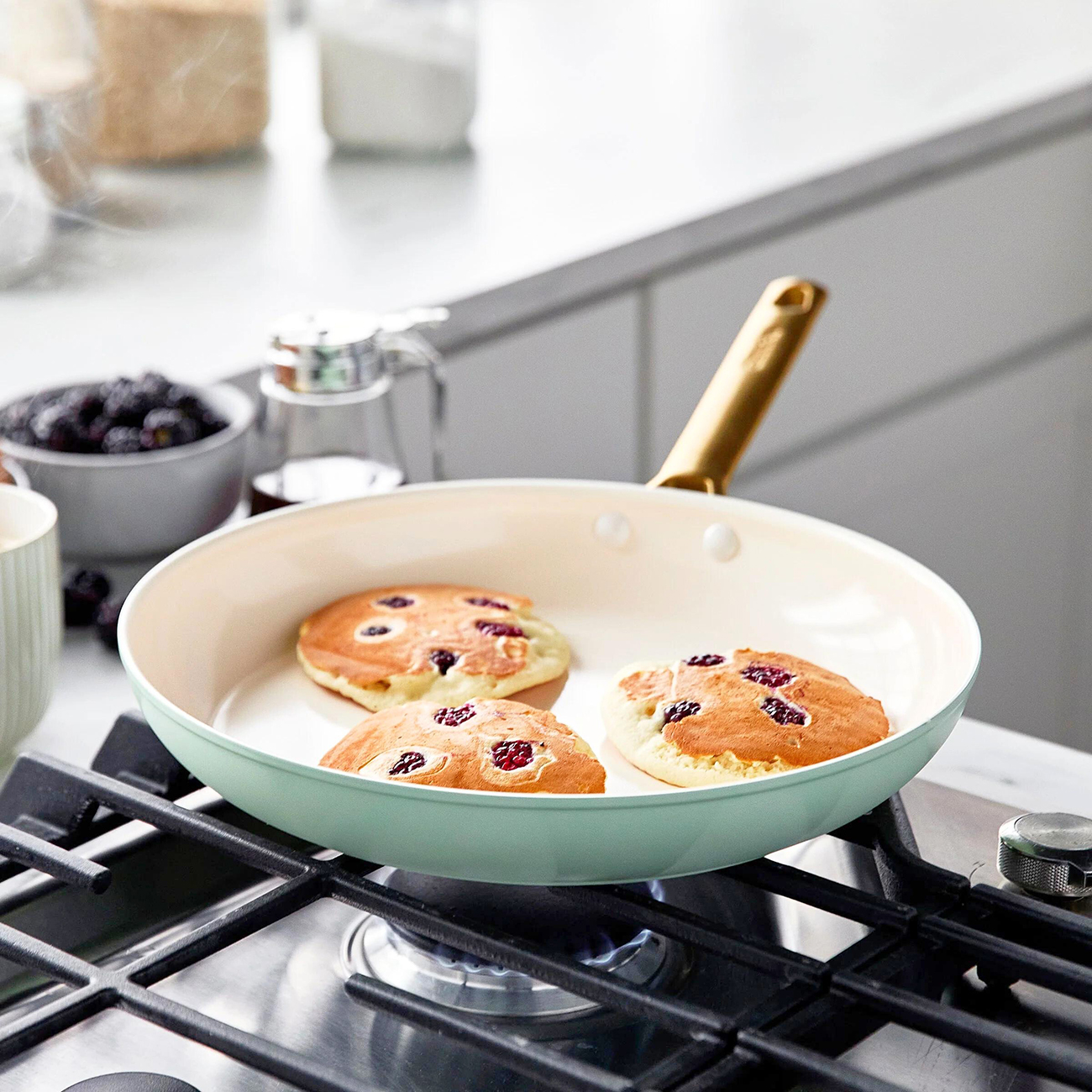 Drei Pfannkuchen mit Beeren werden in einer Pfanne auf einem Gasherd gebraten. Beeren, Sirup und andere Küchenartikel im Hintergrund.