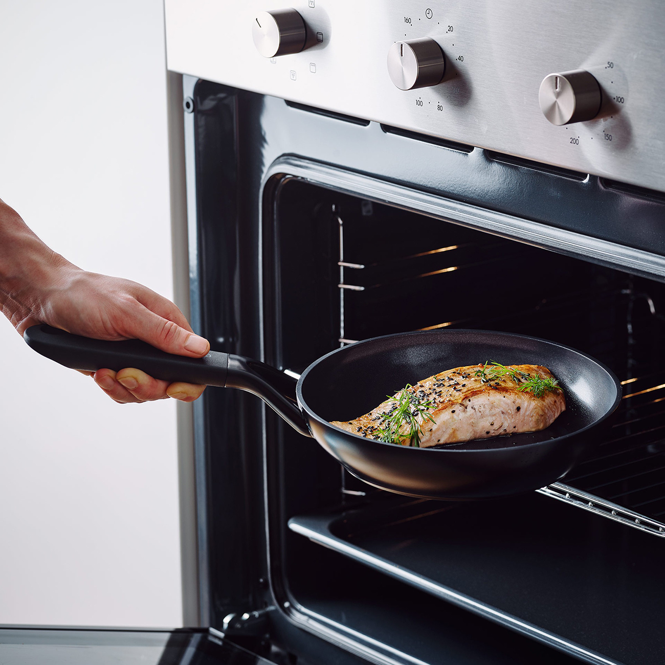 Eine Hand hält eine Pfanne mit gekochtem Lachs und stellt sie in einen Ofen. Der Ofen ist aus Edelstahl mit Drehknöpfen.