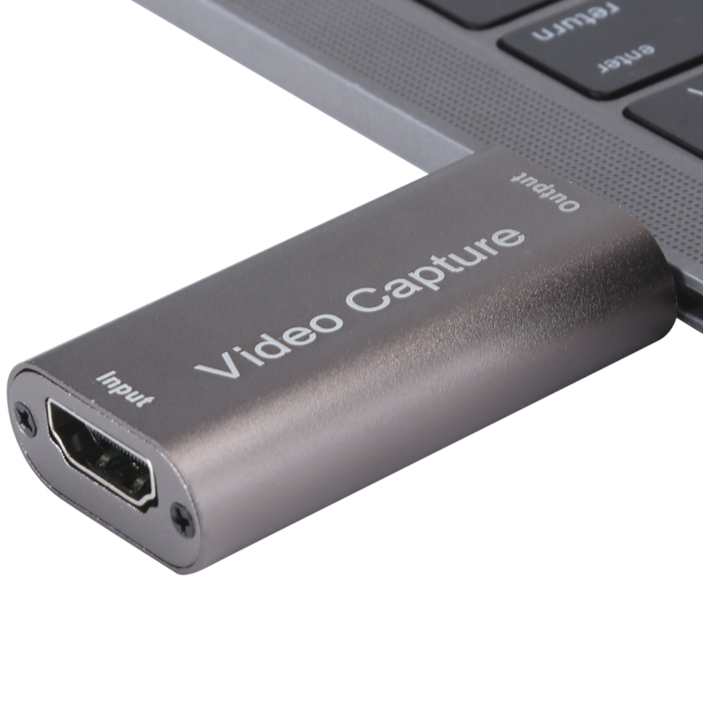 Ein silbernes Videoaufnahmegerät mit der Aufschrift 'Video Capture' ist mit einer Laptop-Tastatur verbunden. Es hat HDMI-Eingangs- und -Ausgangsanschlüsse.