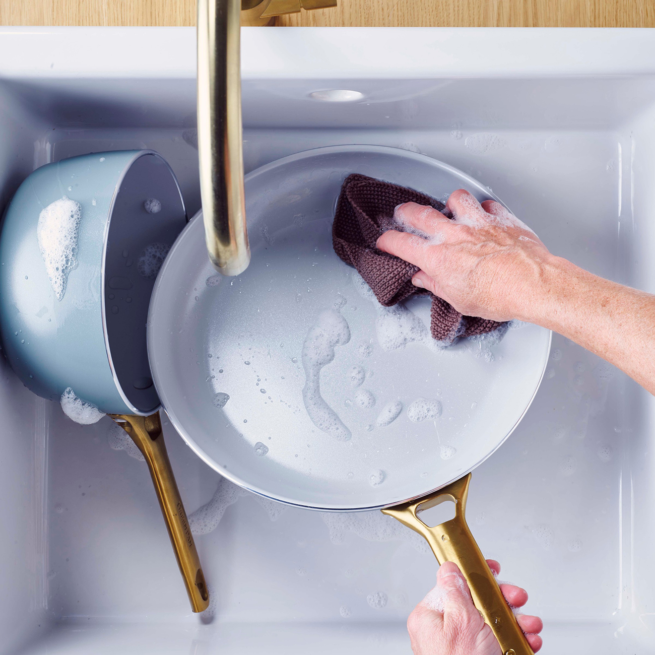 Eine Person wäscht eine Pfanne in einem Waschbecken. Die Pfanne ist hellgrau mit goldenem Griff. Daneben steht ein blauer Topf. Seifenschaum ist reichlich vorhanden.