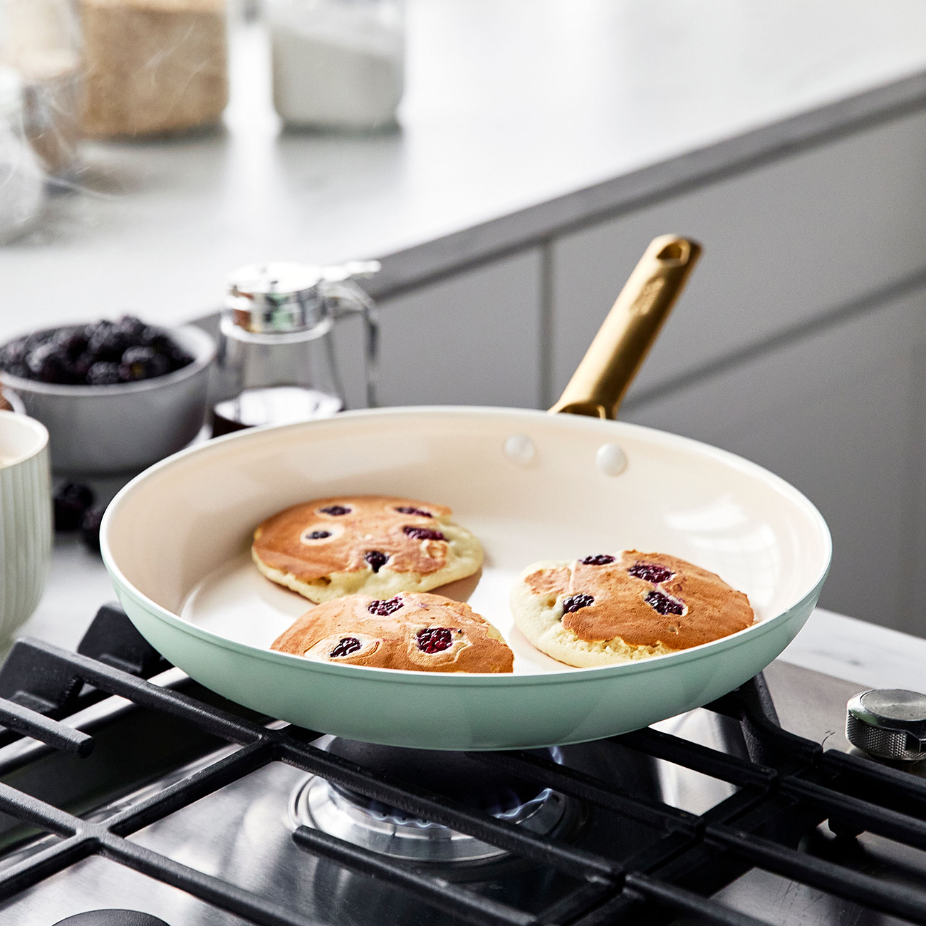 Drei Pfannkuchen mit Beeren werden in einer Pfanne auf einem Herd gebraten. In der Nähe befinden sich eine Schüssel mit Beeren, ein Sirup-Spender und andere Küchenartikel.