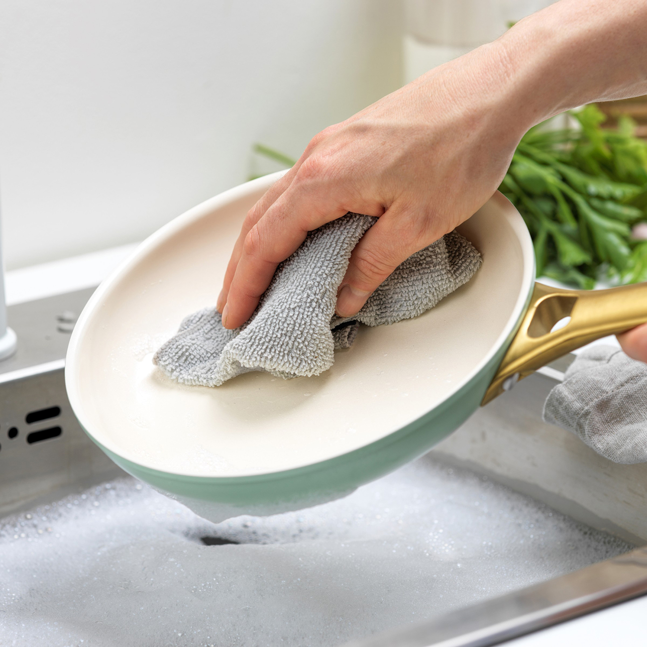 Eine Person wäscht eine Pfanne in einem mit Seifenwasser gefüllten Waschbecken. Die Pfanne ist grün und gold. Ein graues Tuch wird zum Reinigen verwendet.