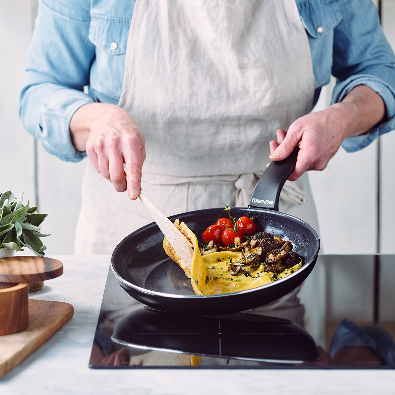 Eine Person kocht in einer Pfanne mit Omelett, Kirschtomaten und Pilzen. Die Person trägt eine Schürze und ein Jeanshemd.