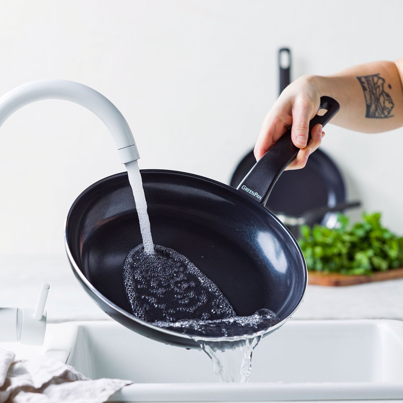 Eine Person wäscht eine schwarze Bratpfanne unter einem weißen Wasserhahn. Wasser gießt in die Pfanne und erzeugt Blasen. Weitere Pfannen und Kräuter sind im Hintergrund.