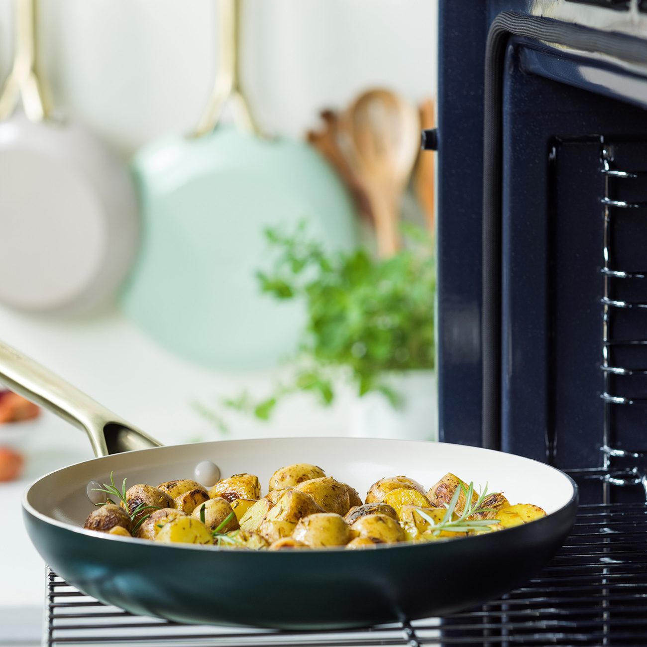Eine Pfanne mit gebratenen Kartoffeln und Rosmarin steht auf einem Rost. Rechts ist ein Ofen. Töpfe hängen im Hintergrund.
