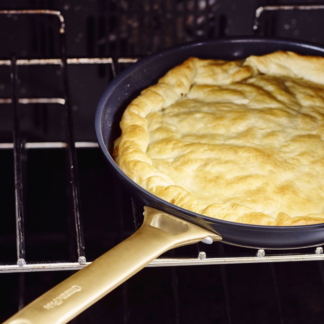 Ein goldbrauner Kuchen in einer dunklen Pfanne im Ofen. Der Griff ist golden. Das Ofengitter ist sichtbar.