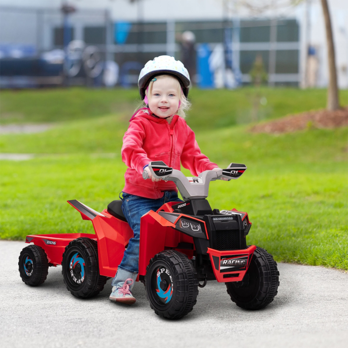 Ein Kind in roter Jacke und Helm fährt auf einem roten Spielzeug-ATV mit Anhänger und lächelt in die Kamera.