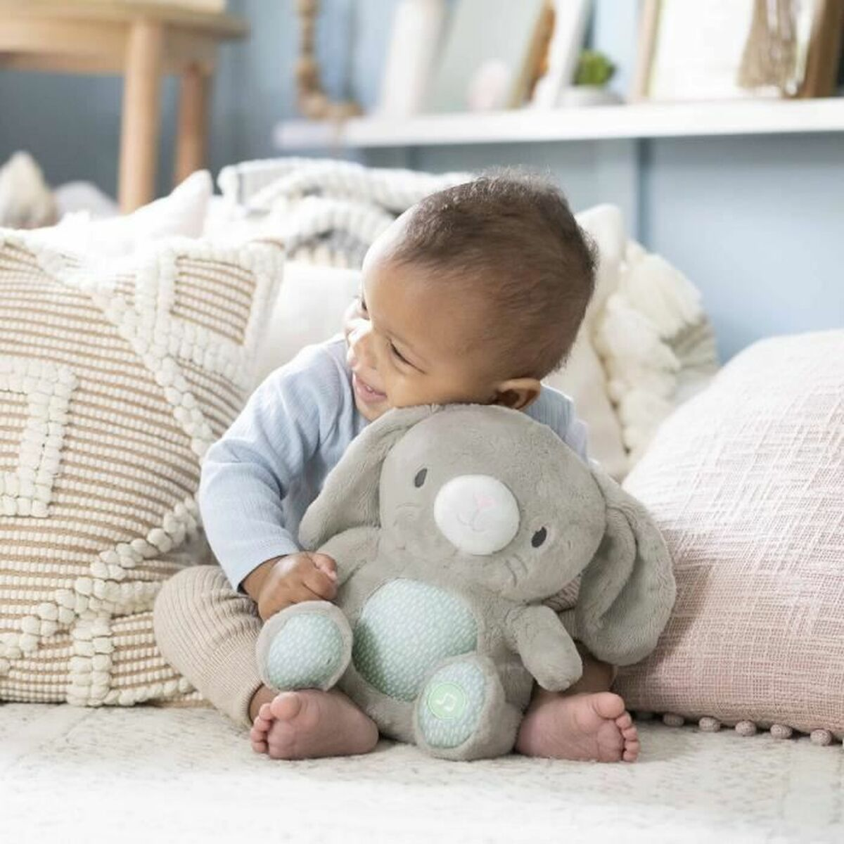 Un bébé sourit en tenant un lapin en peluche gris. Le bébé est assis sur une surface blanche avec des oreillers en arrière-plan.