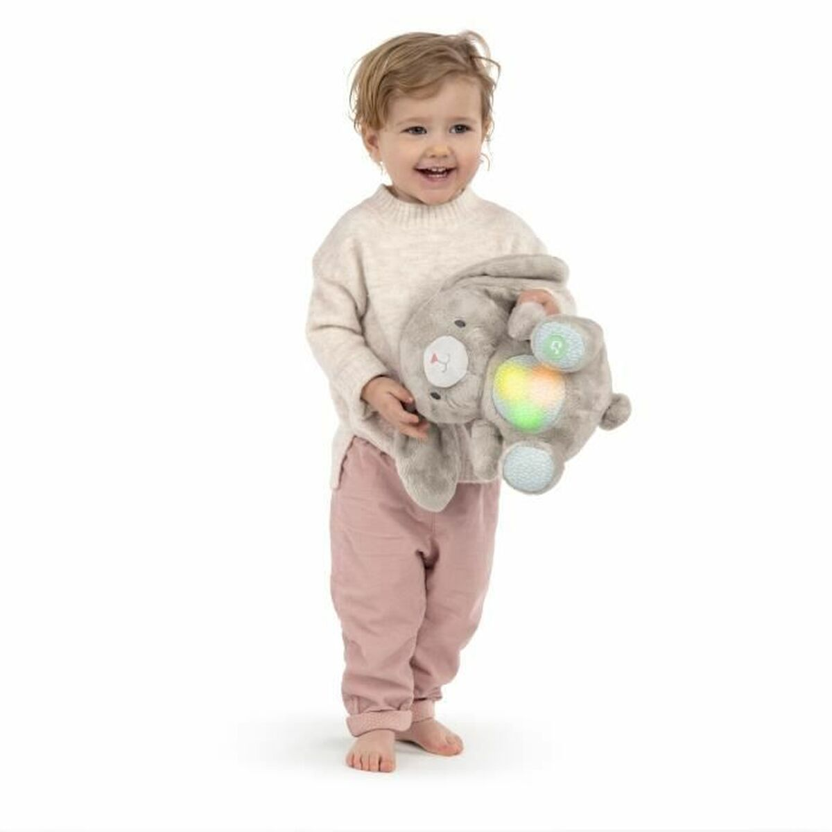 Un tout-petit sourit, tenant un jouet lapin gris avec une lumière arc-en-ciel. Elle porte un pull crème et un pantalon rose, debout pieds nus.