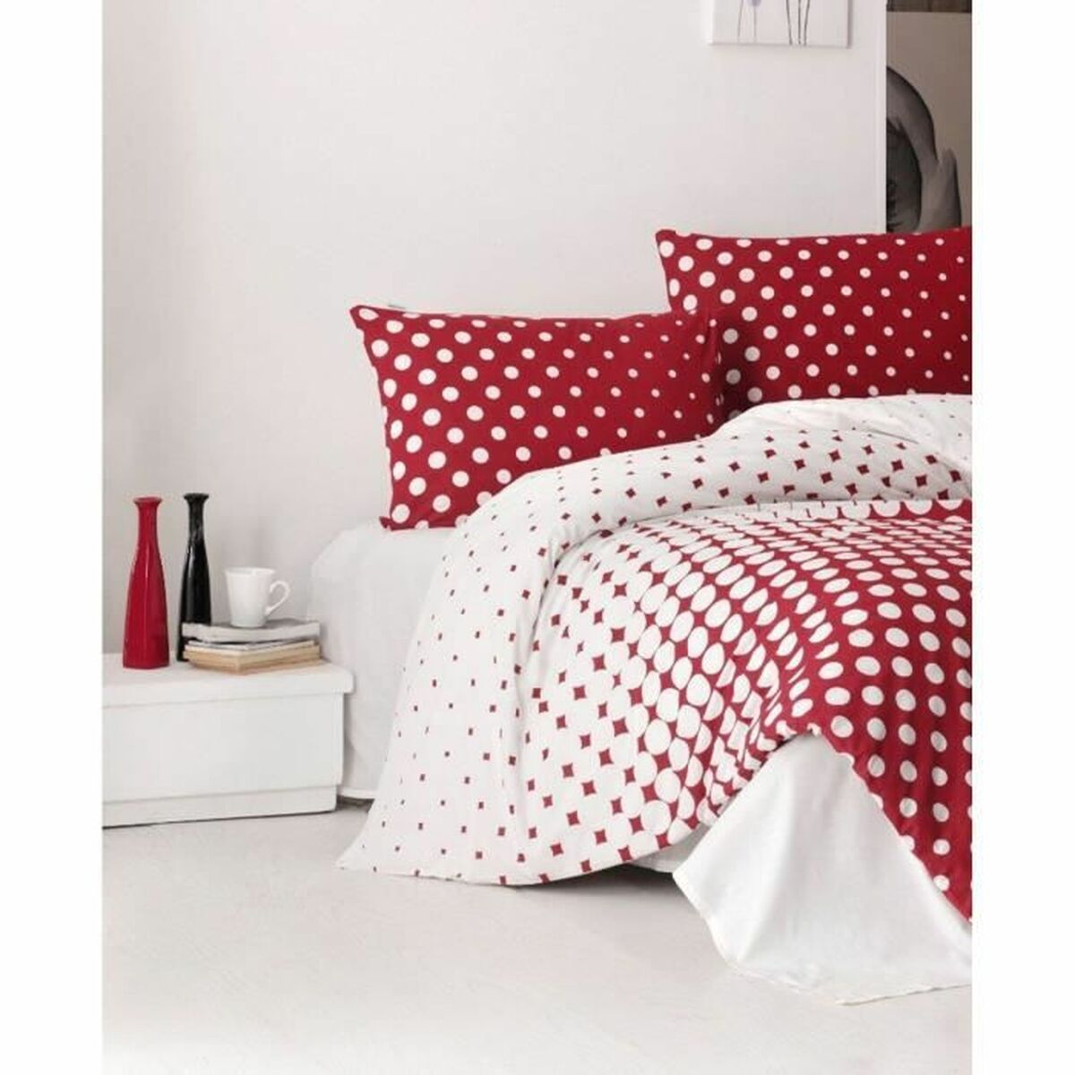 Rote und weiße Polka-Dot-Bettwäsche auf einem Bett. Zwei rote Vasen und eine Tasse stehen auf einem weißen Nachttisch. Weiße Wand.