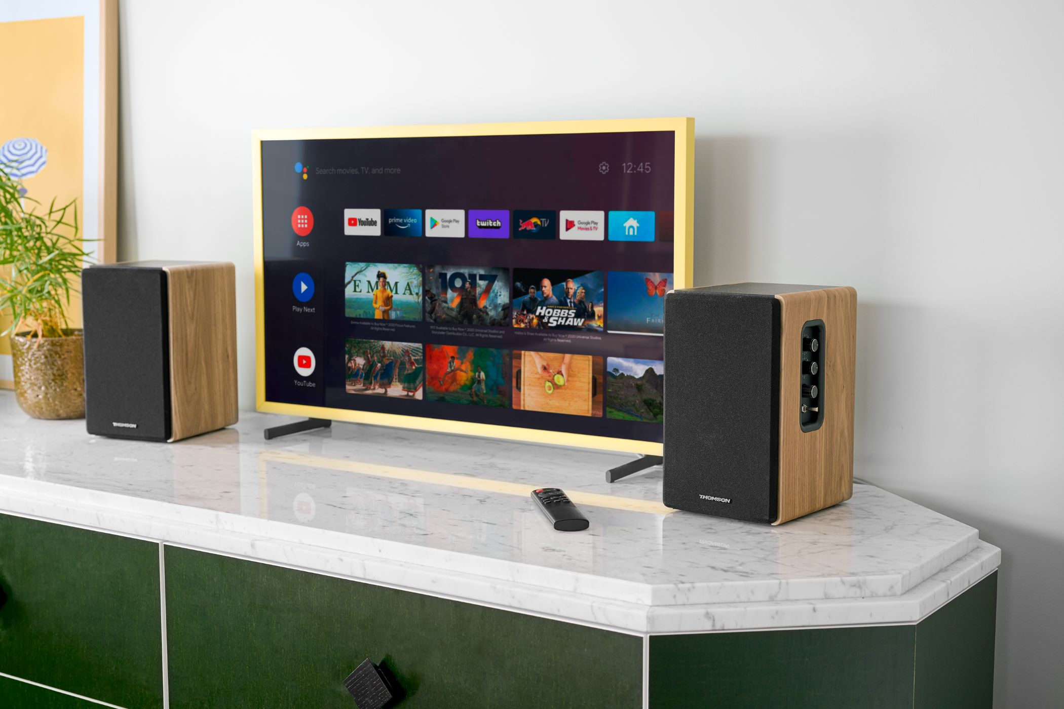 Telewizor wyświetla aplikacje do strumieniowania i wybór filmów. Głośniki flankują telewizor na białej marmurowej szafce. Pilot znajduje się na szafce.