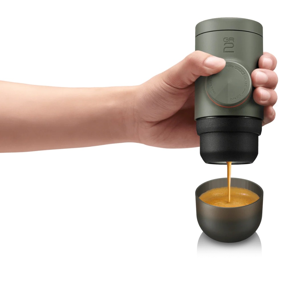 Eine Hand hält einen tragbaren Espressomacher und gießt Kaffee in eine kleine Tasse. Das Gerät ist grün und schwarz, die Tasse ist grau.
