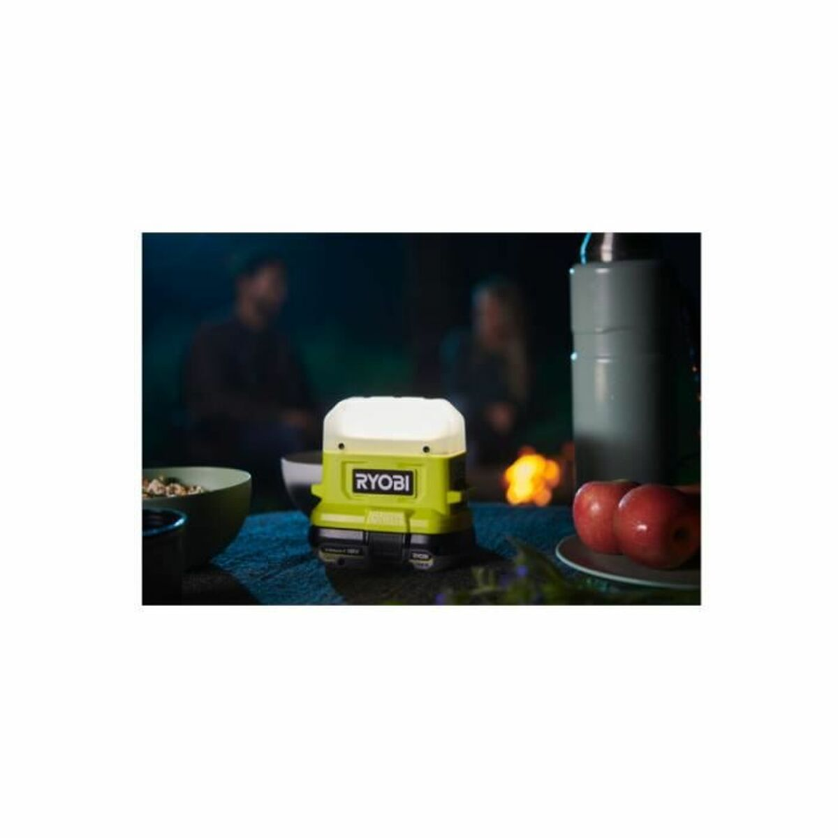 Eine grüne Ryobi-Lampe auf einem blauen Tisch. Äpfel, eine Thermoskanne und eine Schüssel befinden sich in der Nähe. Menschen und ein Feuer sind im Hintergrund.
