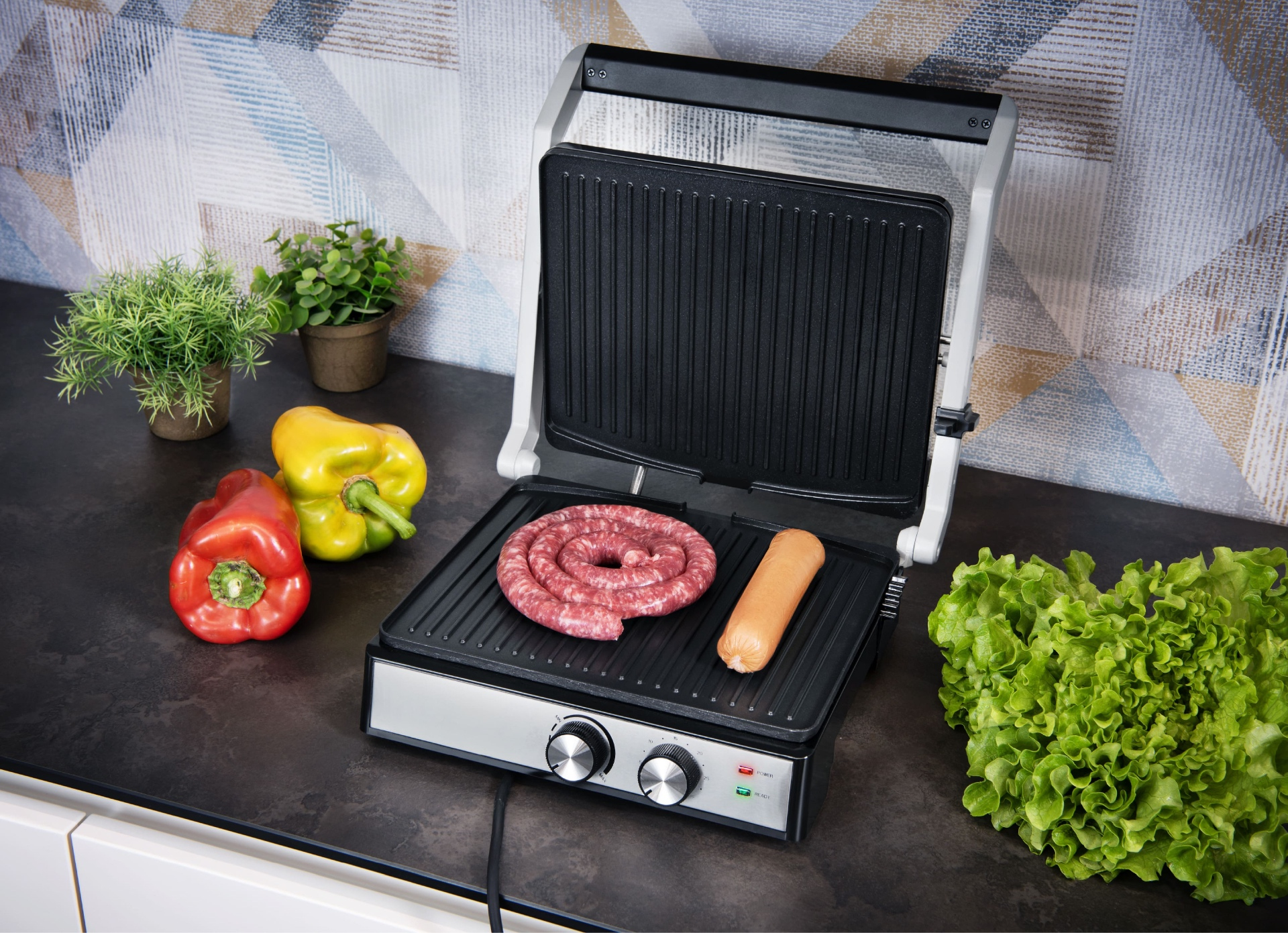 Grill z kiełbasą i zwiniętą kiełbasą na ciemnej powierzchni. Widoczne są również papryka, sałata i rośliny.