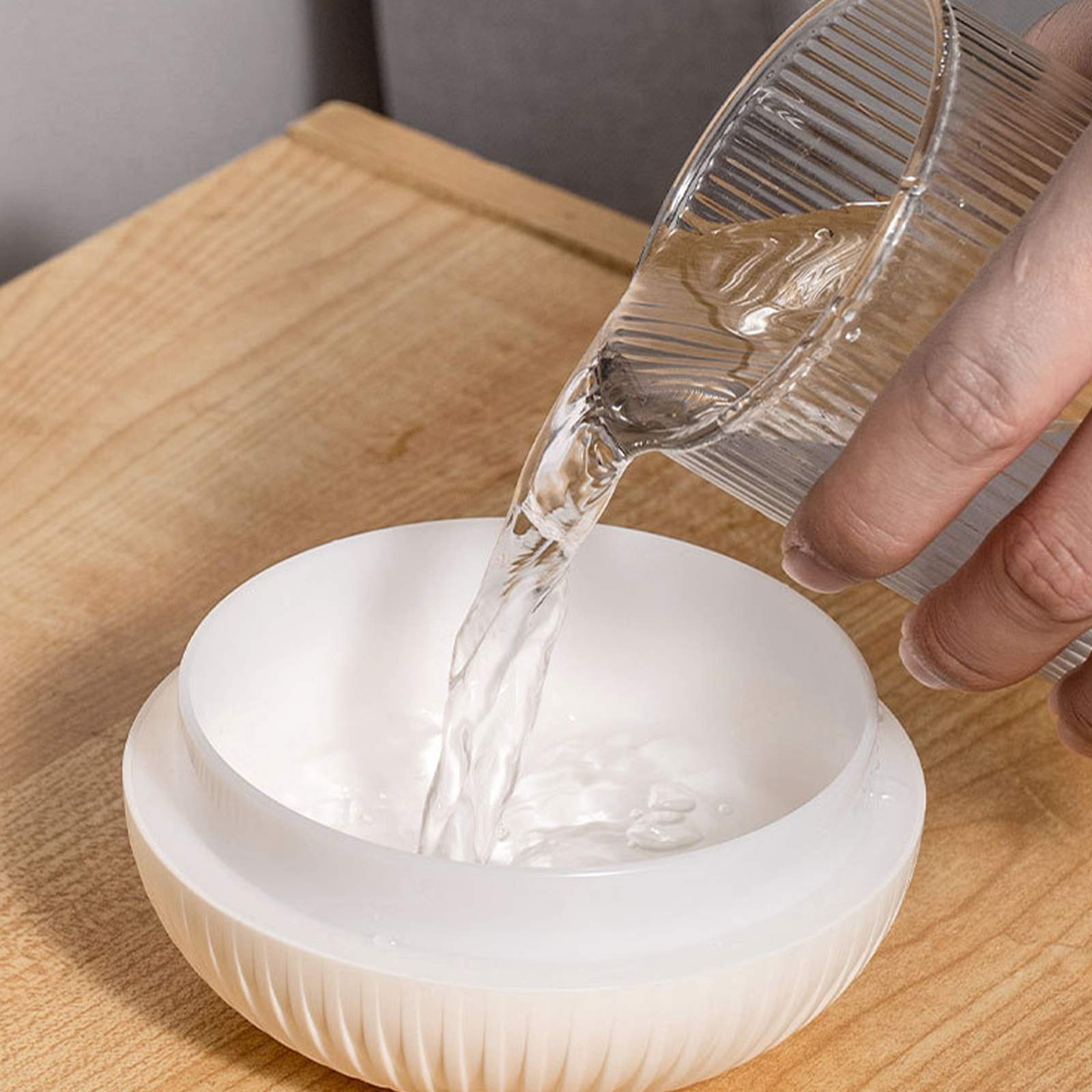 Wasser wird aus einem geriffelten Glas in eine weiße Schüssel auf einer Holzoberfläche gegossen. Die Hand hält das Glas.