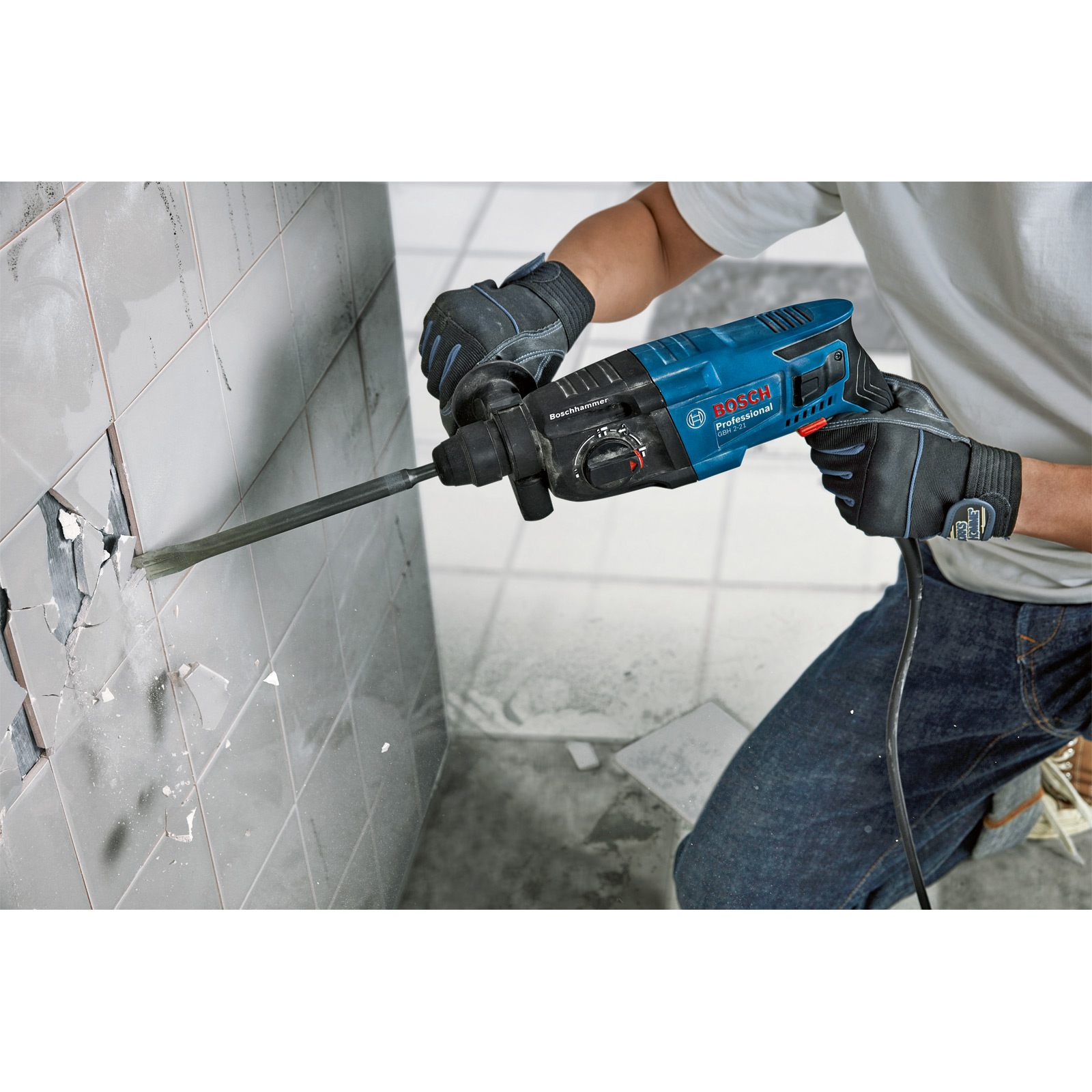 Eine Person benutzt einen blauen Bosch-Hammerbohrer, um Fliesen an einer Wand zu zerbrechen. Die Wand ist beschädigt, die Person trägt Handschuhe.