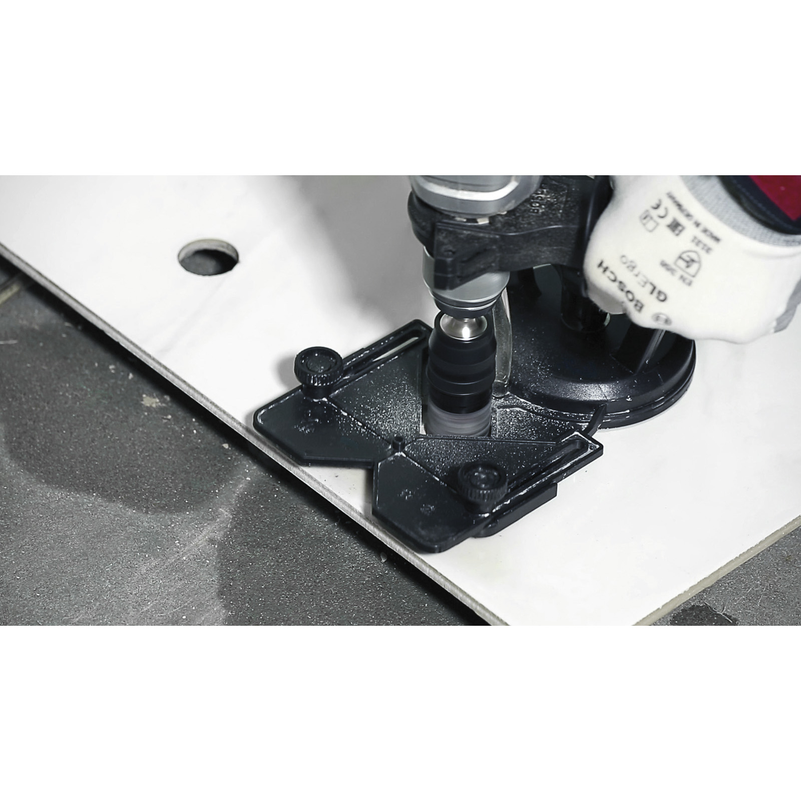 Eine Bohrmaschine wird auf einer weißen Fliese verwendet. Eine schwarze Führung ist angebracht. Eine behandschuhte Hand ist sichtbar.